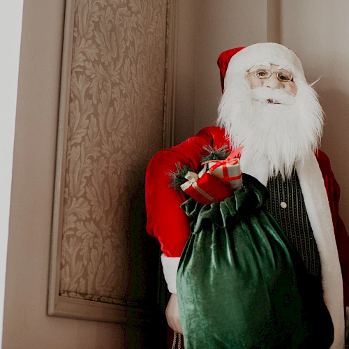 Santa Claus in a red suit carrying a green sack, standing in a doorway with beige walls and patterned paneling.