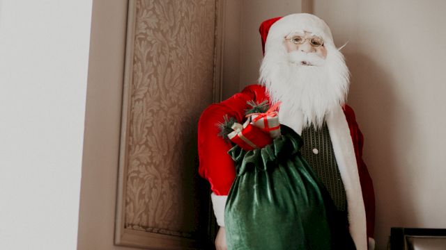 Santa Claus in a red suit carrying a green sack, standing in a doorway with beige walls and patterned paneling.