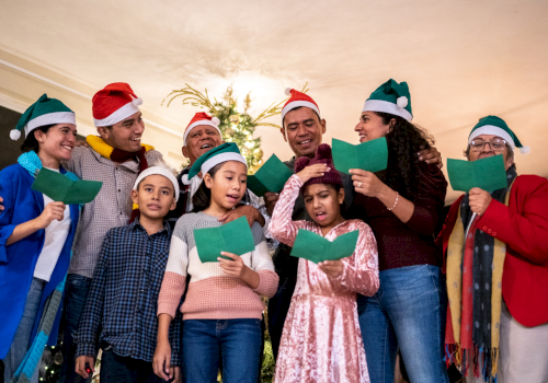 A diverse group sings carols indoors, wearing festive hats and holding green songbooks, gathered around a Christmas tree.