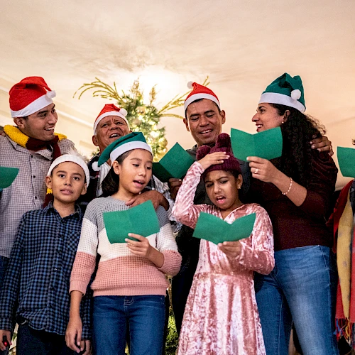 A diverse group sings carols indoors, wearing festive hats and holding green songbooks, gathered around a Christmas tree.