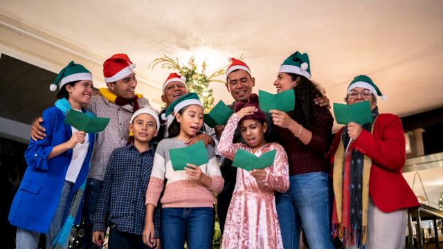 A diverse group sings carols indoors, wearing festive hats and holding green songbooks, gathered around a Christmas tree.