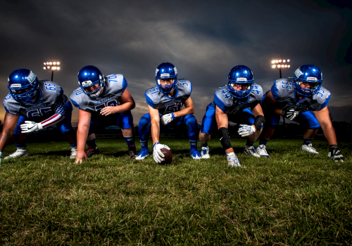 A football offensive line crouches in a three-point stance on a grassy field under stadium lights at dusk, ready for the snap.