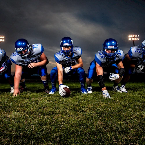 A football offensive line crouches in a three-point stance on a grassy field under stadium lights at dusk, ready for the snap.
