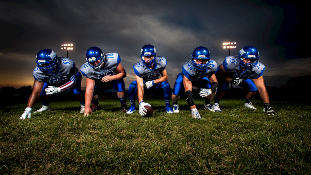A football offensive line crouches in a three-point stance on a grassy field under stadium lights at dusk, ready for the snap.