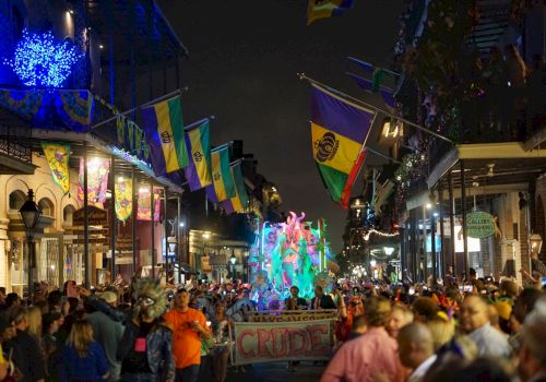 A crowded nighttime parade fills a street with colorful flags, bright lights, and a glowing float waving through the festival.
