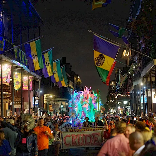 A crowded nighttime parade fills a street with colorful flags, bright lights, and a glowing float waving through the festival.