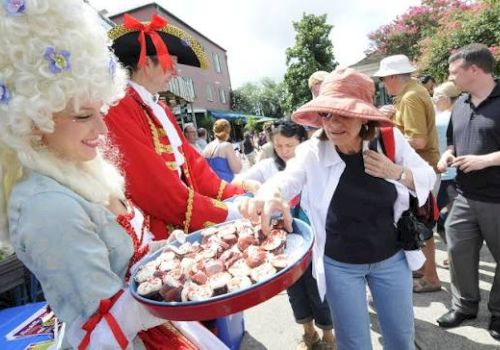 A street festival shows costumed performers offering strawberries from a tray to passersby in a sunny crowd.