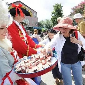 A street festival shows costumed performers offering strawberries from a tray to passersby in a sunny crowd.