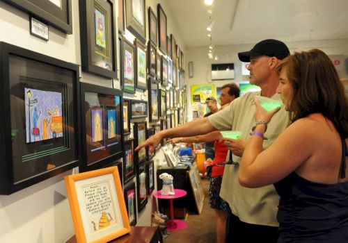 A couple and others browse framed artworks in a hallway gallery, pointing while viewing colorful paintings on the walls.