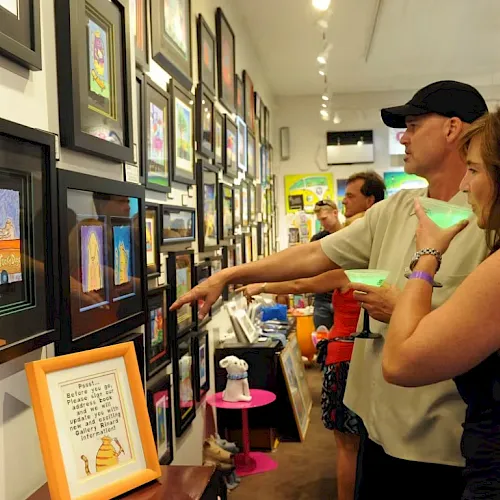 A couple and others browse framed artworks in a hallway gallery, pointing while viewing colorful paintings on the walls.