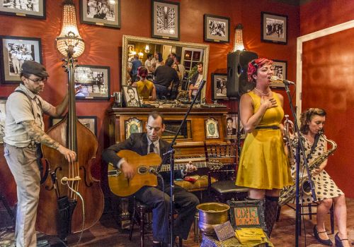A small band performs indoors&mdash;upright bassist, guitarist/singer, and two women singing/playing for an audience in a cozy room.