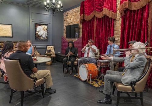 A small jazz group plays brass and drums in a lounge with red curtains while an audience sits and watches.