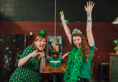 Two women in green St. Patrick&rsquo;s outfits cheer with hands up in a cozy bar, one wearing a green hat and garland, celebrating loudly.
