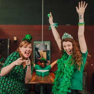 Two women in green St. Patrick’s outfits cheer with hands up in a cozy bar, one wearing a green hat and garland, celebrating loudly.