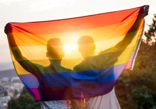 A rainbow Pride flag is held up by two people above a sunset city view.