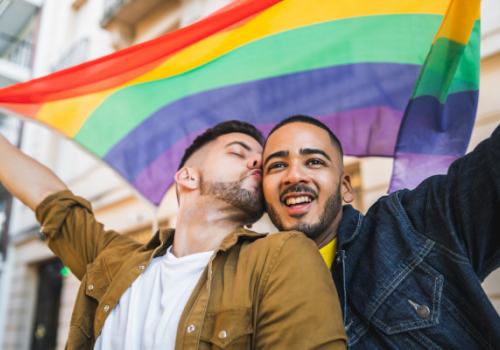 Two men celebrate under a rainbow Pride flag, one kissing the other&rsquo;s cheek while they smile and raise their arms in joy.