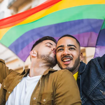 Two men celebrate under a rainbow Pride flag, one kissing the other’s cheek while they smile and raise their arms in joy.