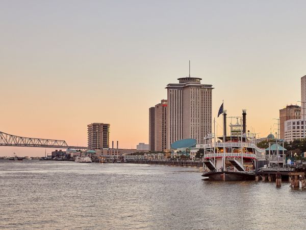 A riverside city skyline with a docked paddleboat and a tall government-style tower, plus a steel bridge in the distance at sunset.