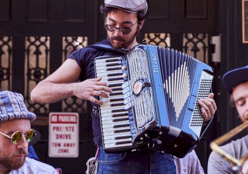 A street musician in a cap plays a blue accordion outdoors while others sit nearby at a public venue.