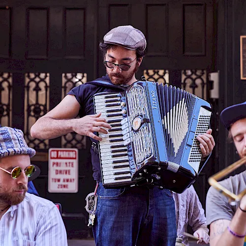 A street musician in a cap plays a blue accordion outdoors while others sit nearby at a public venue.