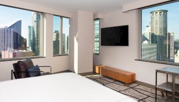 A modern hotel room with city views, featuring large windows, a TV, a bed, a chair, and a small table.
