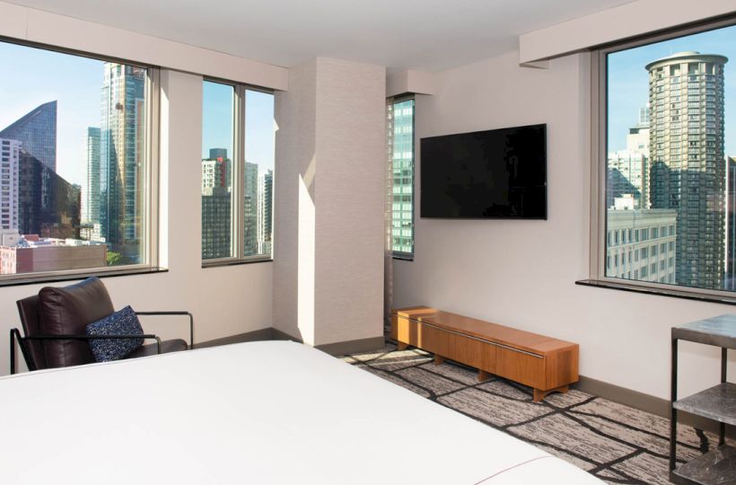 A modern hotel room with city views, featuring large windows, a TV, a bed, a chair, and a small table.