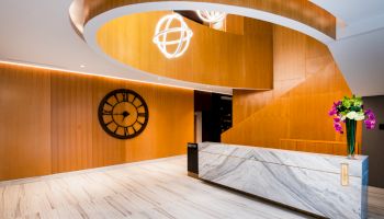 A modern lobby with wooden walls, a marble reception desk, decorative lights, a clock on the wall, and a vase of flowers.