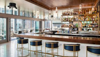 A modern bar interior with a long countertop, stylish blue stools, and shelves stocked with various bottles in a bright, sunlit space.