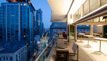 A modern balcony overlooks a city street with tall buildings, featuring tables and chairs in an urban evening setting.