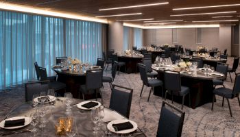 A banquet room with round tables set for an event, featuring black chairs, white flowers, and elegant glassware under soft lighting.