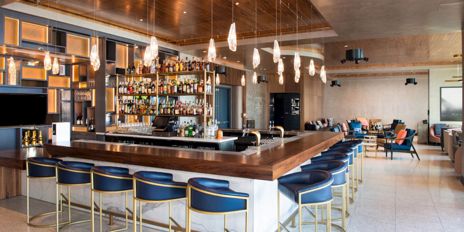 The image shows a modern bar with a central counter, blue stools, hanging lights, and a well-stocked liquor display.