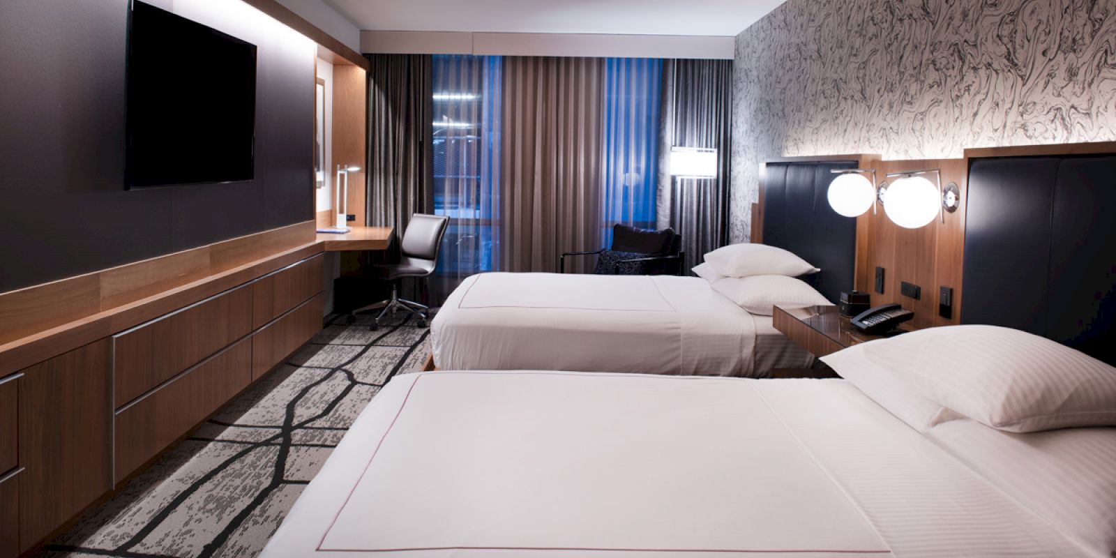 The image shows a modern hotel room with two beds, a TV, desk, chair, and patterned wallpaper, all in neutral tones.