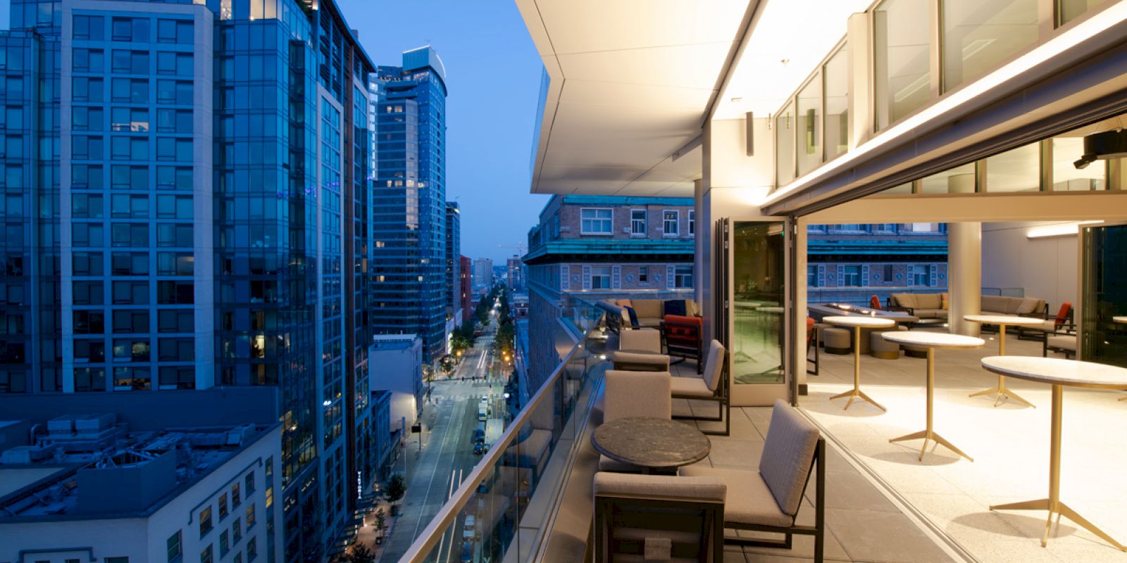 A modern rooftop terrace with seating overlooks a cityscape of tall buildings at dusk, featuring an illuminated, open indoor space.