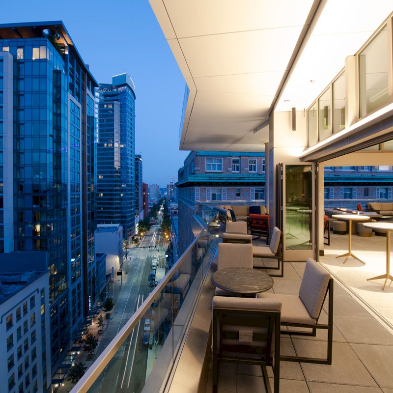 A cityscape view from a modern balcony during evening, with city lights, glass railings, and outdoor seating visible on the terrace.
