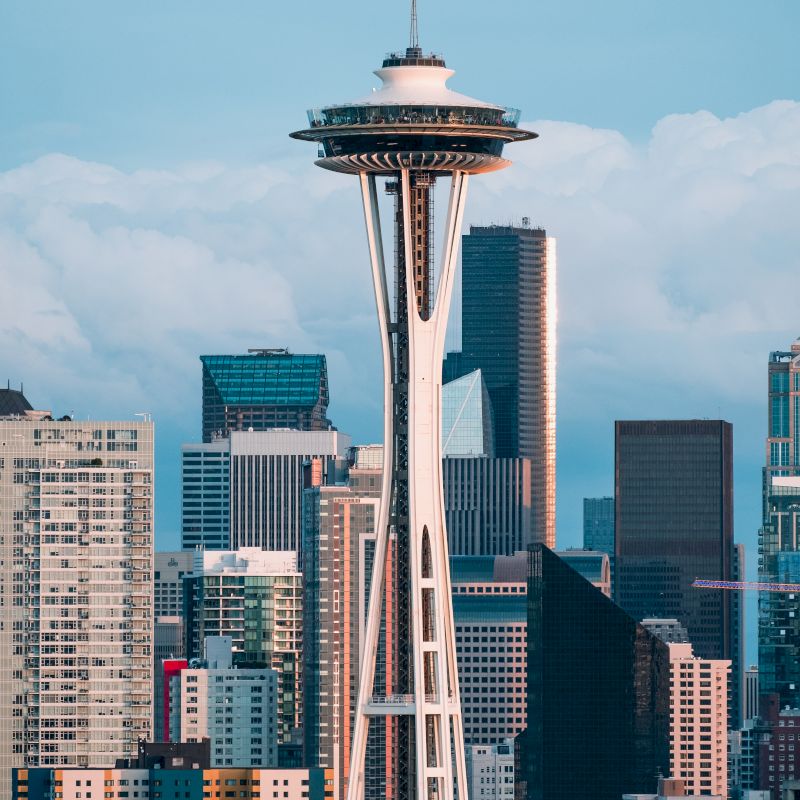 The image shows a skyline with a prominent tower, likely the Space Needle, surrounded by various modern buildings and skyscrapers under a blue sky.