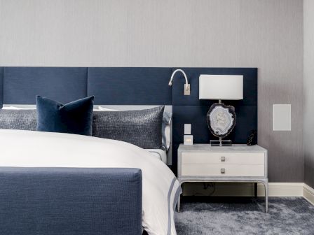 A modern bedroom with a blue upholstered headboard, white linens, and a nightstand holding a lamp and decorative object.