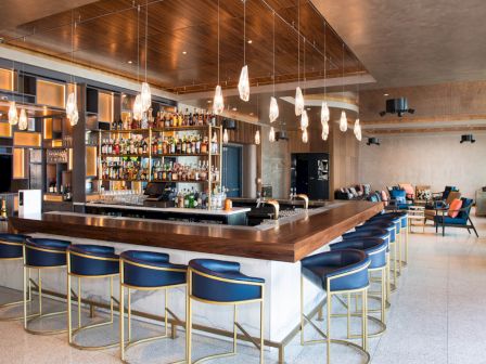 A modern bar with stylish blue stools, a well-stocked liquor display, pendant lights, and a lounge area with seating in the background.