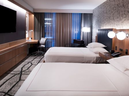 The image shows a modern hotel room with two beds, a desk, a TV, and stylish lighting, featuring a patterned carpet and wall decor.