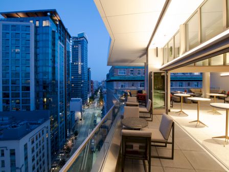 A modern cityscape view from a balcony at dusk, featuring buildings, a street below, and outdoor seating with tables and chairs.
