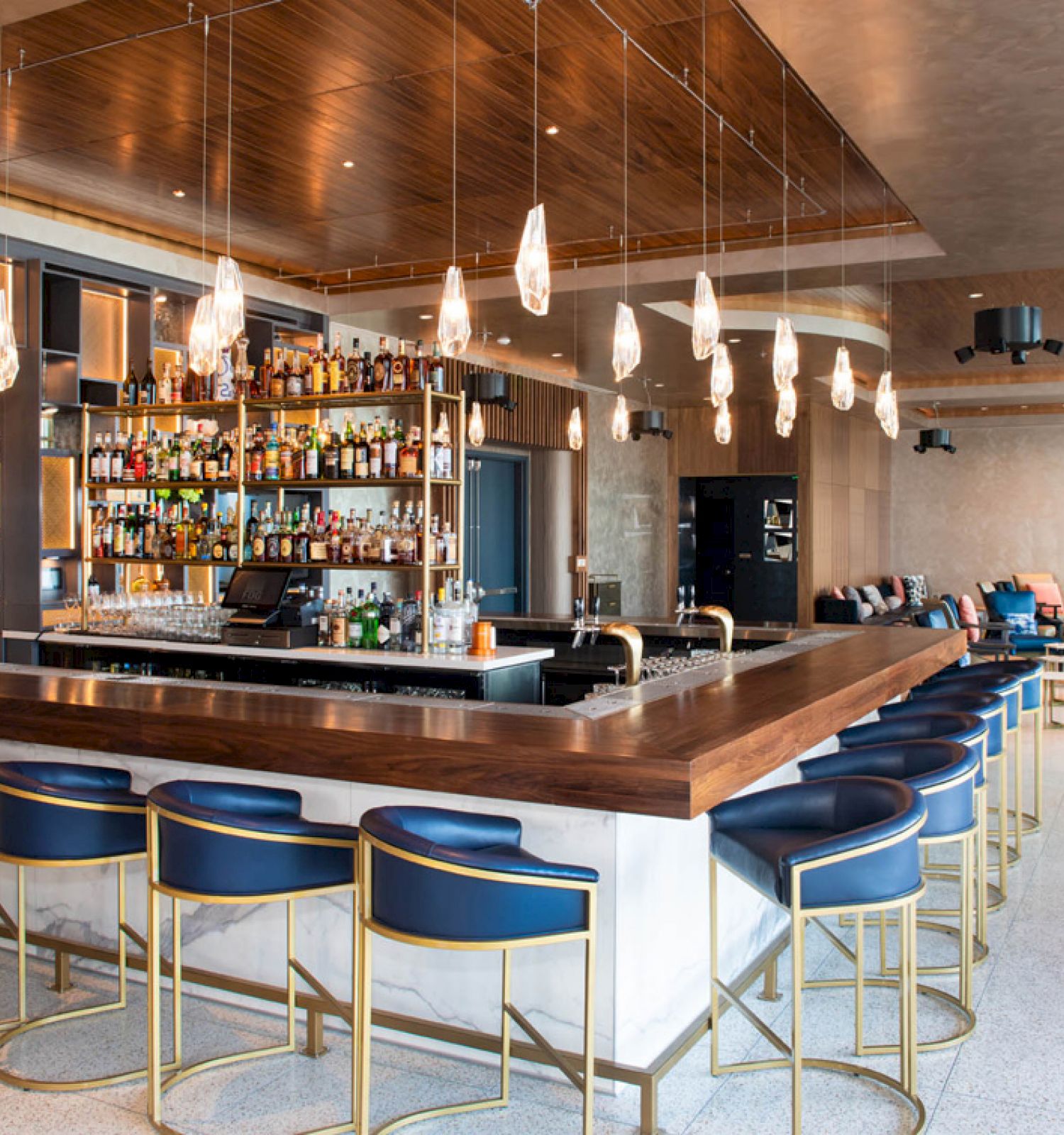 A modern bar with blue chairs, a wooden counter, and pendant lights. Shelves are stocked with bottles, seating area visible in the background.