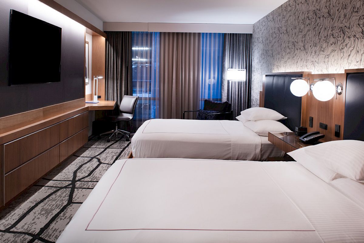 The image shows a modern hotel room with two beds, a TV, a desk, and stylish lighting. The room has a contemporary design and decor.