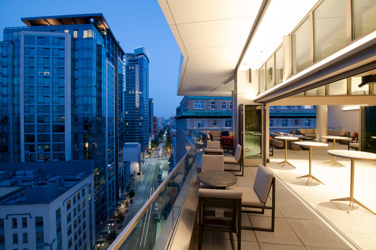 A modern balcony view of a city skyline at dusk, featuring outdoor seating and illuminated buildings lining the street.