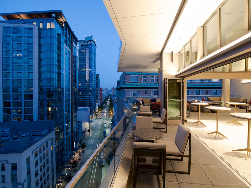 A modern balcony view of a city skyline at dusk, featuring outdoor seating and illuminated buildings lining the street.