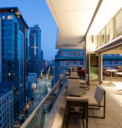 A modern balcony overlooks a cityscape at dusk, featuring tables and seating, with nearby tall buildings and a street view.