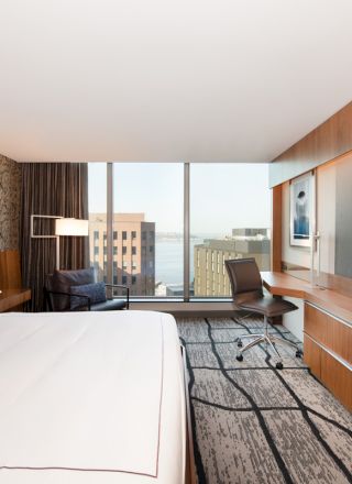 A modern hotel room features a bed, desk, chair, large TV, and window with a city view. The decor is sleek and contemporary.