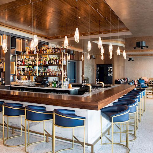 A modern bar with stylish lighting, a full bottle display, and blue cushioned chairs creating a sleek, inviting atmosphere.