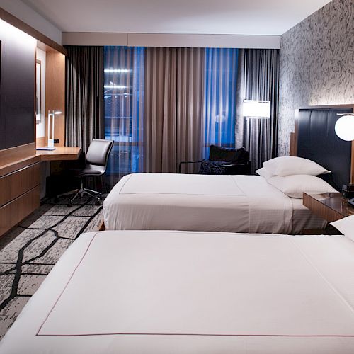 The image shows a modern hotel room with two beds, wall-mounted TV, desk, chair, and stylish decor.