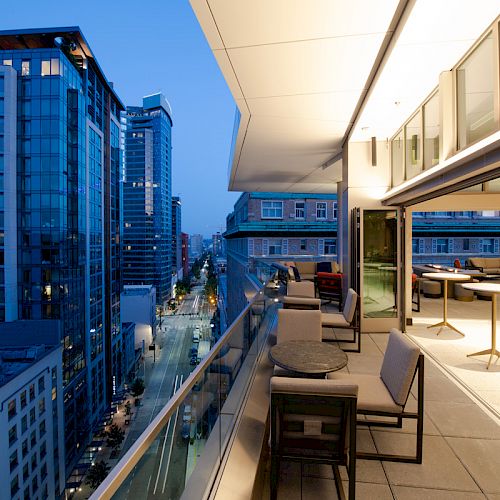 The image shows a modern cityscape at dusk with lit buildings and a stylish outdoor terrace with seating and tables.