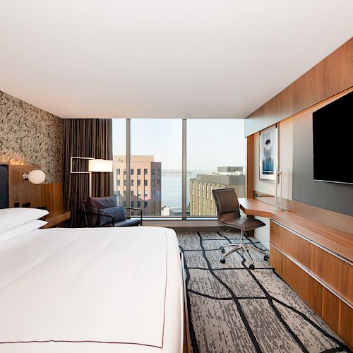 A modern hotel room with a large bed, desk, chair, and a TV on the opposite wall, featuring a city view through large windows.