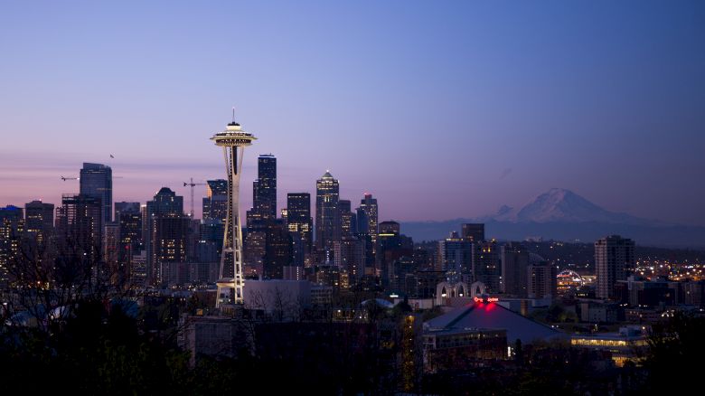 The image shows a city skyline at dusk with the Space Needle prominently visible and a mountain in the background.